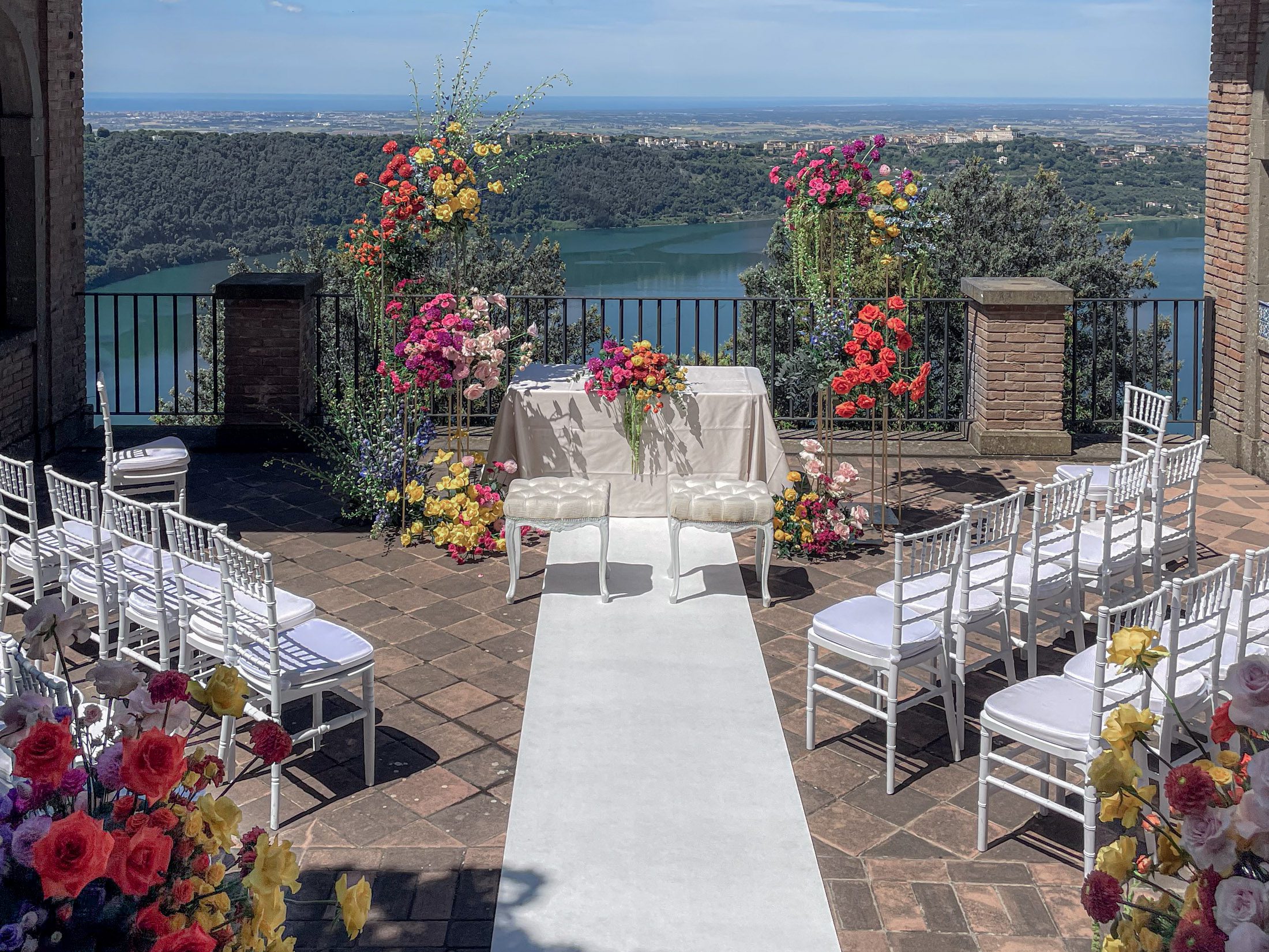 Cerimonia di matrimonio all’aperto con vista panoramica su un lago, decorata con fiori vivaci e sedie bianche – allestimento floreale firmato Inanna Event